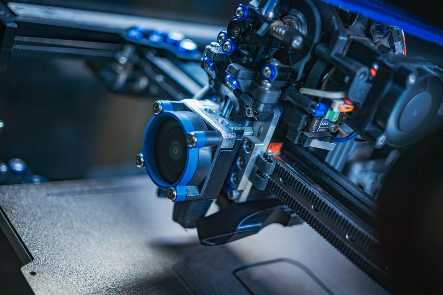 Primer plano detallado de la cinemática de una impresora 3D, mostrando el extrusor, las correas dentadas y los rieles lineales.