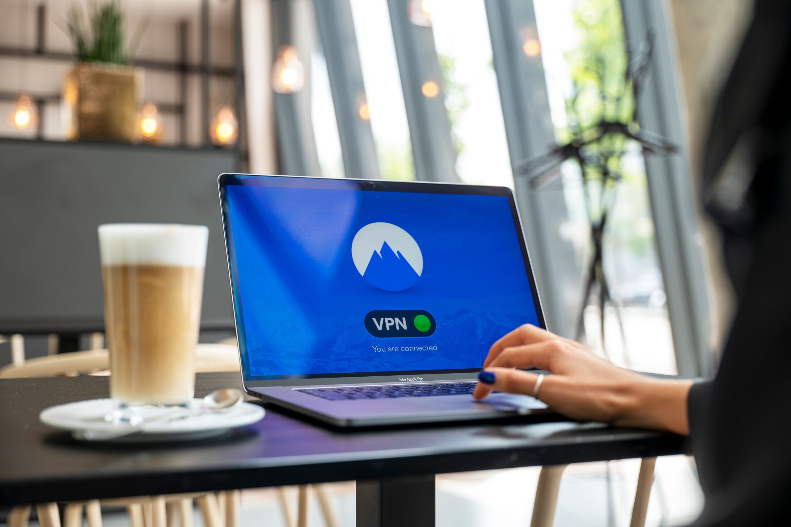 Persona utilizando un ordenador portátil en una cafetería moderna con la conexión VPN activada para navegar de forma segura en redes Wi-Fi públicas.