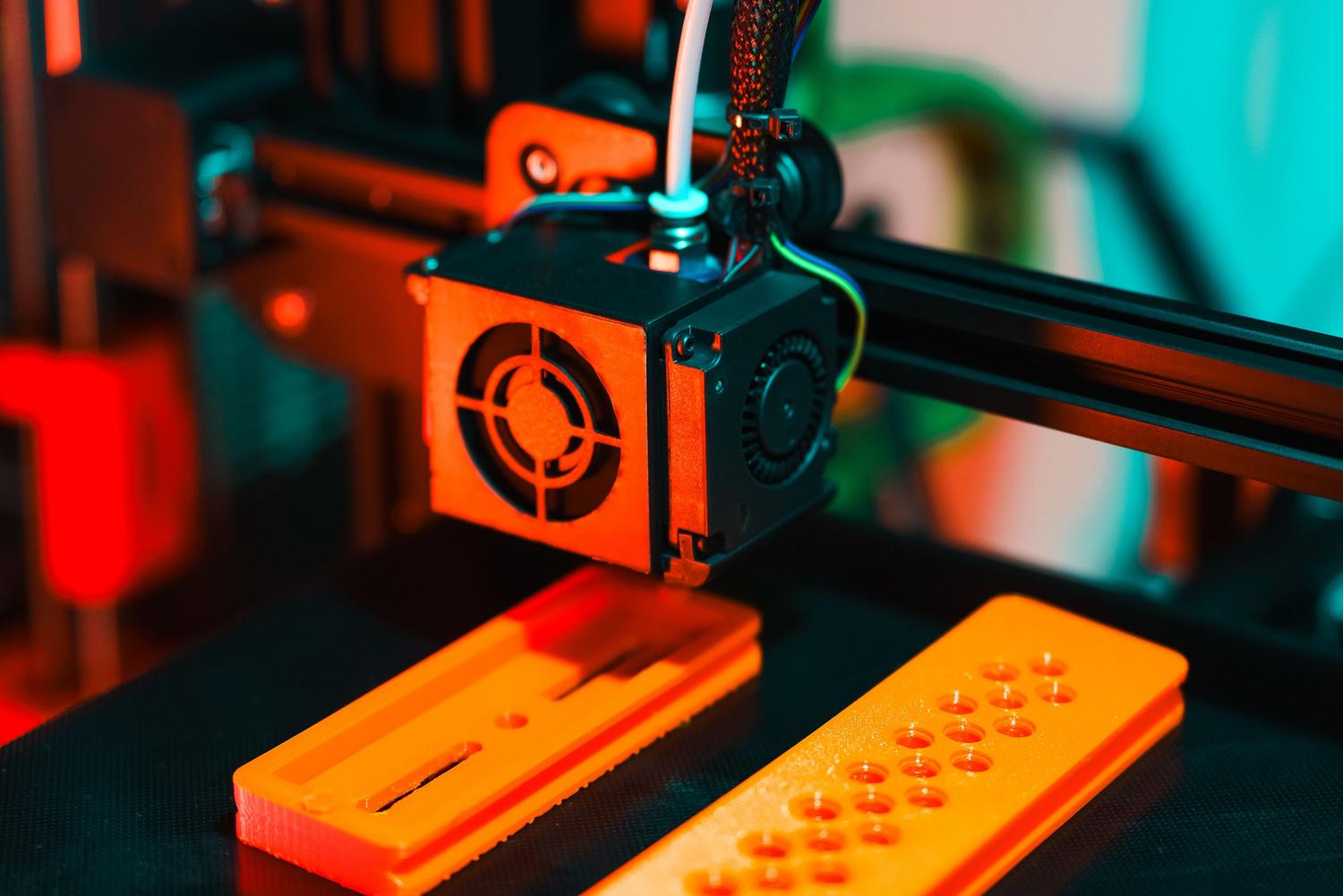Primer plano de una boquilla de impresora 3D extruyendo filamento naranja, ilustrando el funcionamiento de la producción aditiva.