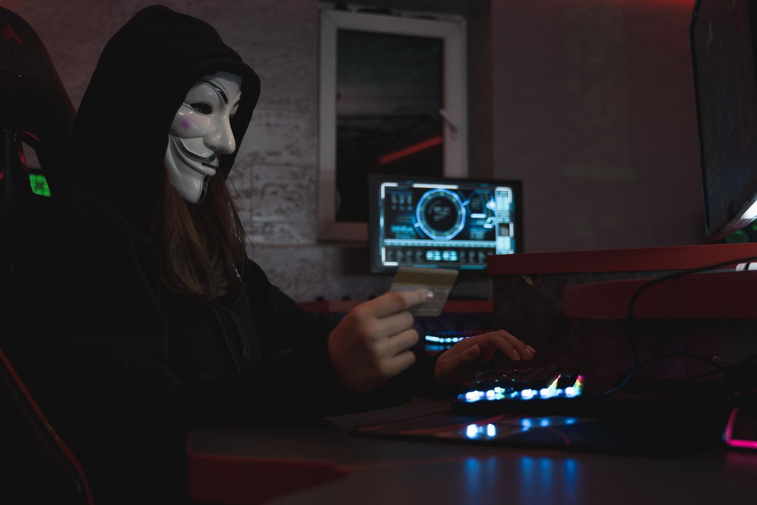 Hacker enmascarado sosteniendo una tarjeta de crédito frente a un ordenador, ilustrando las operaciones de cibercrimen.