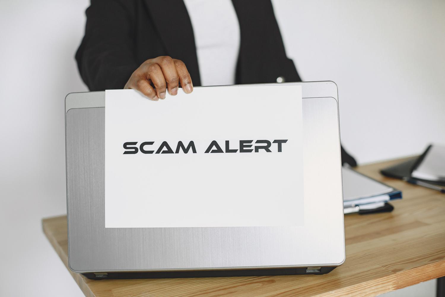 Persona sosteniendo un cartel de alerta de estafa sobre un ordenador portátil, advirtiendo sobre el fraude online.