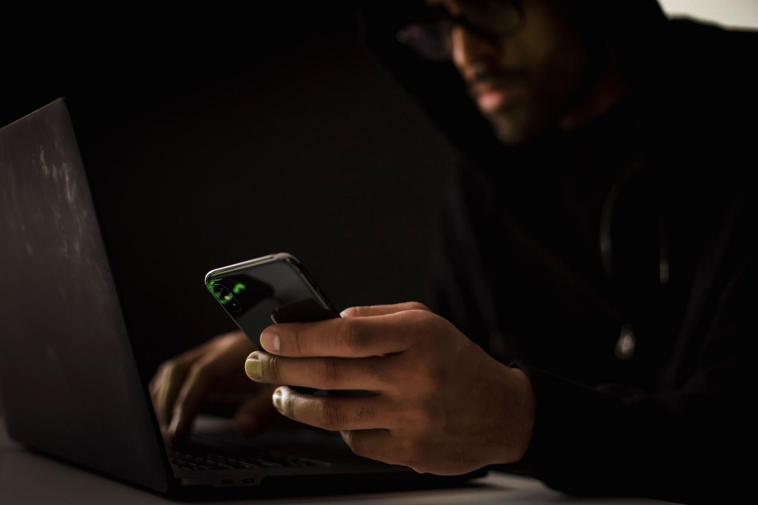 Ciberdelincuente utilizando un ordenador y un móvil en la oscuridad para representar la amenaza de los hackers y la ingeniería social