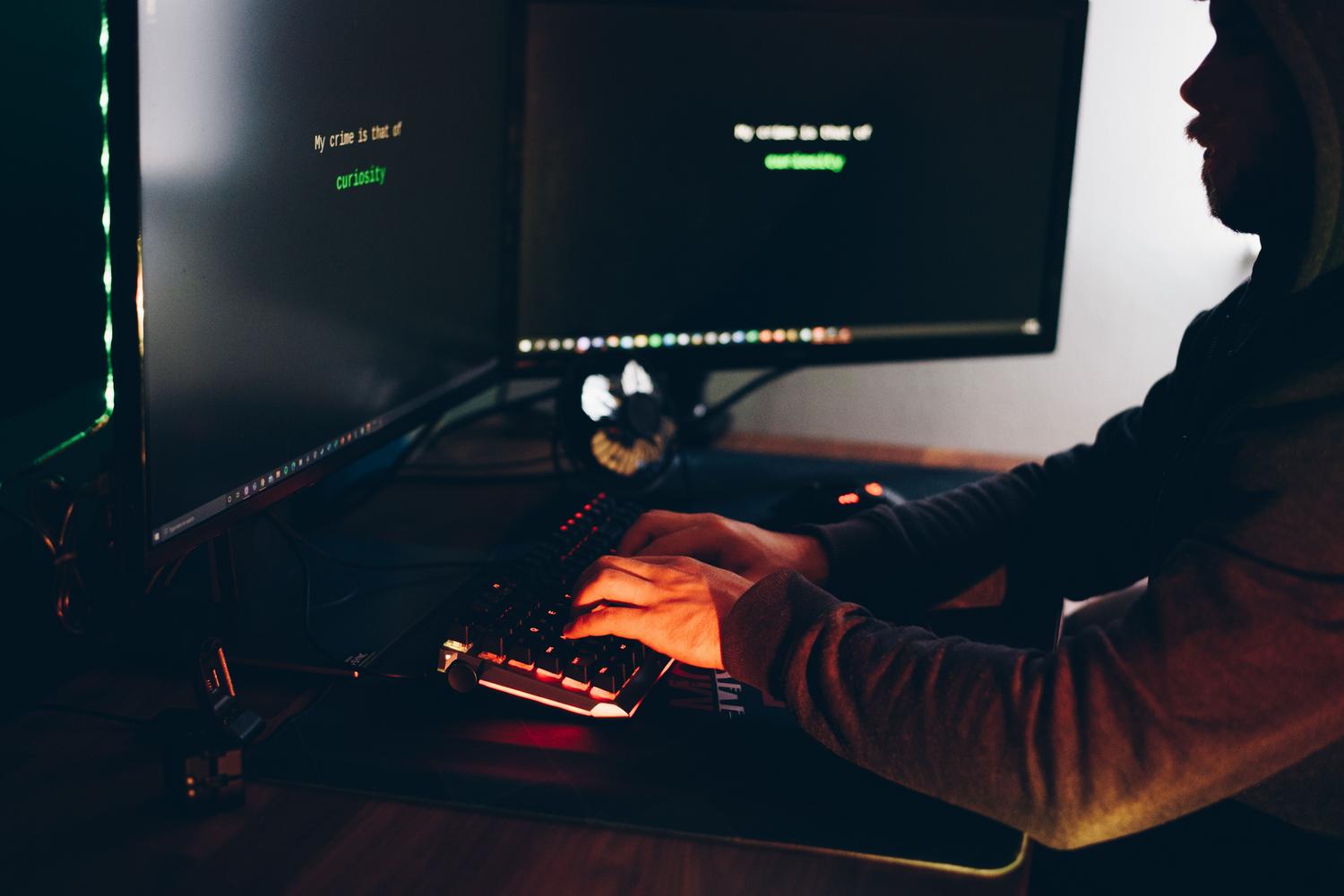 Silueta de un hacker encapuchado operando una computadora en la penumbra, simbolizando la amenaza externa y la explotación de vectores de ataque.
