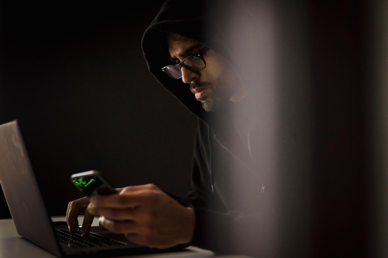 Ciberdelincuente con capucha utilizando un smartphone y un portátil en un entorno oscuro, representando la ingeniería social y el phishing.
