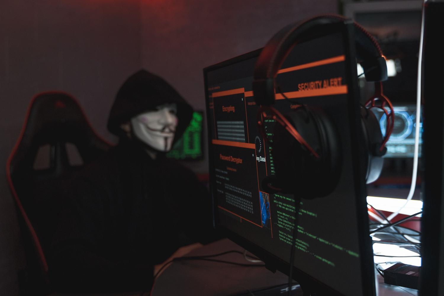 Persona encapuchada realizando actividades de hacking en un ordenador en una habitación oscura, simbolizando la amenaza de los ciberdelincuentes.