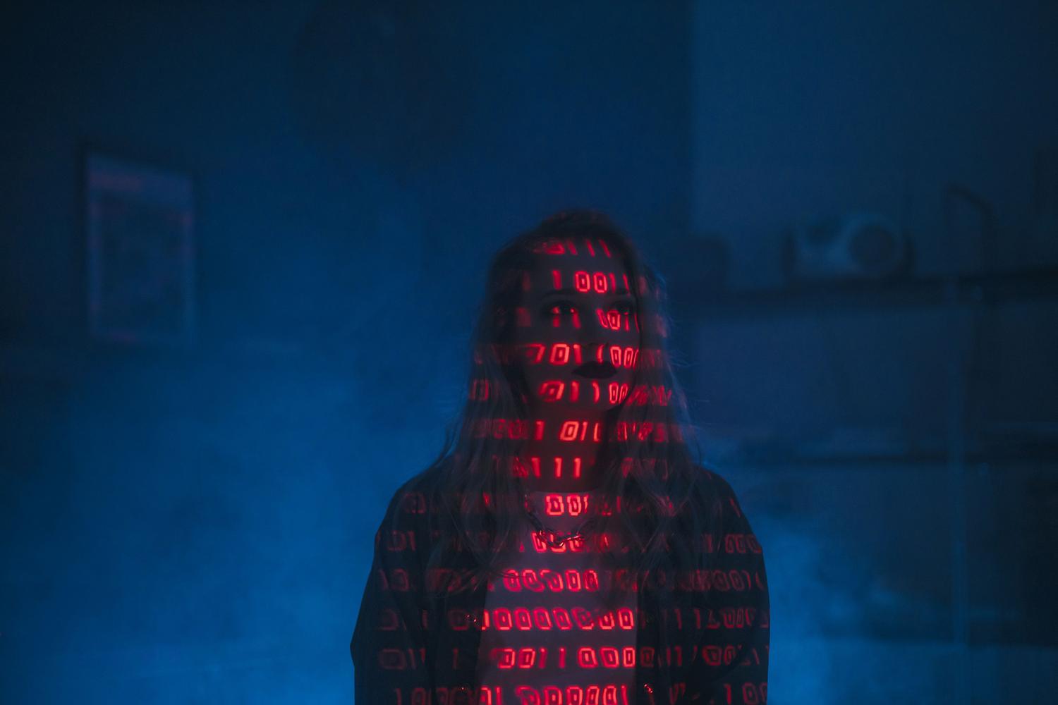 Retrato conceptual de una persona con código binario rojo y azul proyectado sobre su rostro, simbolizando el anonimato de los ciberdelincuentes y la manipulación de datos.