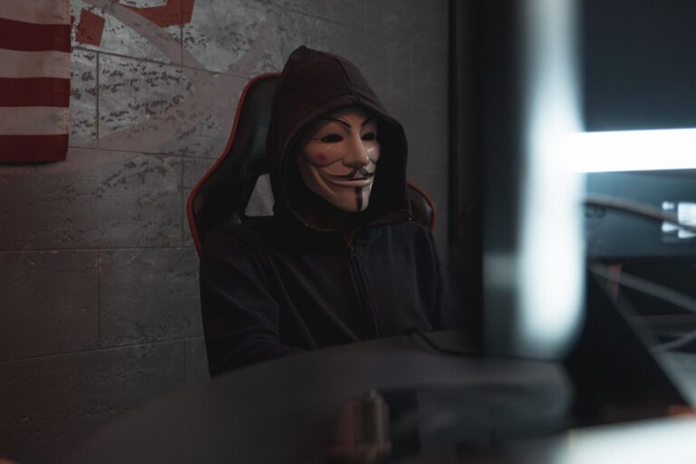 Ciberdelincuente con máscara de Guy Fawkes y capucha frente a un ordenador en un entorno oscuro, representando el anonimato de los hackers.
