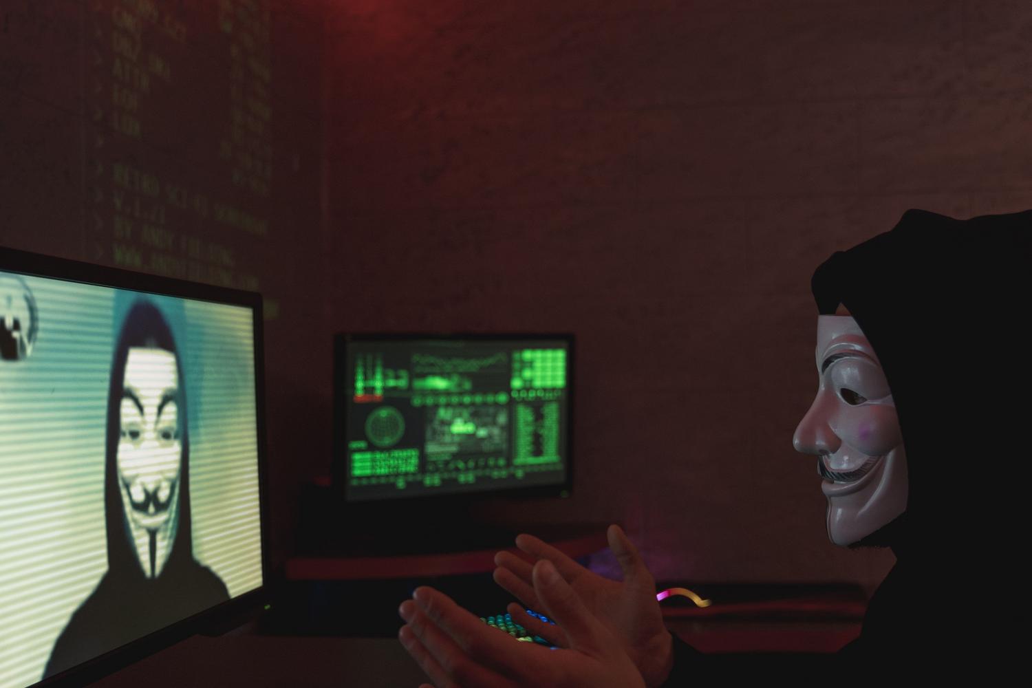 Ciberdelincuente anónimo con máscara de Guy Fawkes coordinando un ataque digital desde la oscuridad frente a monitores con código.